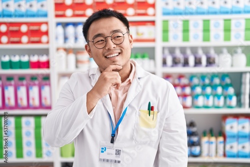 Chinese young man working at pharmacy drugstore with hand on chin thinking about question, pensive expression. smiling and thoughtful face. doubt concept. © Krakenimages.com