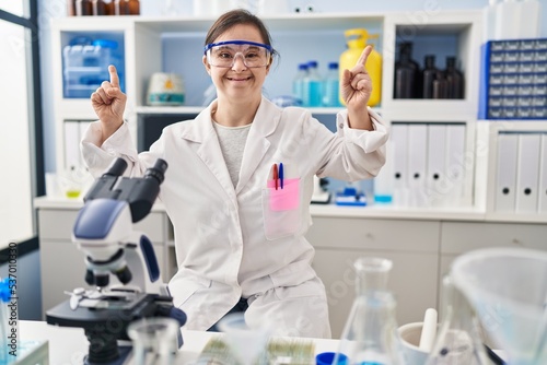 Hispanic girl with down syndrome working at scientist laboratory smiling amazed and surprised and pointing up with fingers and raised arms.