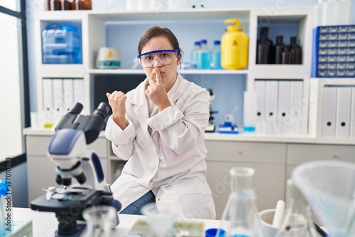 Hispanic girl with down syndrome working at scientist laboratory asking to be quiet with finger on lips pointing with hand to the side. silence and secret concept.