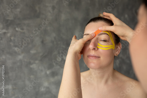 Cropped photo of therapist doctor woman applying orange yellow kinesio tapes on forehead of middle-aged woman patient. photo