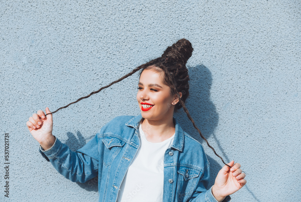 A brunette girl with dreadlocks and red lipstick, in a denim suit, is standing near a blue wall. generation z A brunette girl with dreadlocks and red lipstick, in a denim suit, is standing near a blue wall. generation z