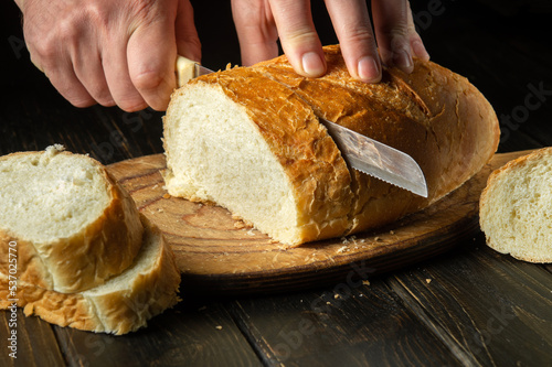 Slicing freshly baked wheat bread with a knife by the hands of a chef. National dish on the kitchen table