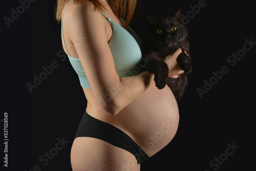 A beautiful pregnat woman with black cat on black background, Motherhood. Fashion photo for pregnancy, Big pregnant belly.