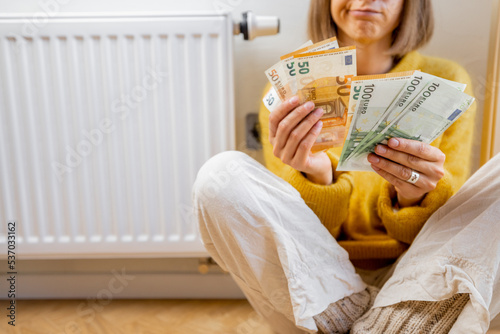 Warmly dressed woman counting money while sitting near radiator at home. Concept of expensive energy resources and the energy crisis in Europe