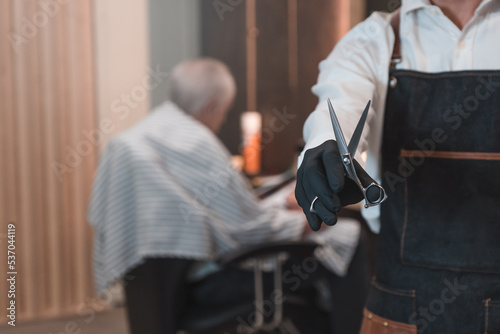 Detalle de peluquero irreconocible, enseñando las tijeras y de fondo cliente senior, leyendo una revista a la espera de cortarse el pelo. © Ezequiel Martínez