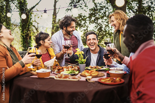 Company of happy people chatting dining at the barbecue table relaxing together eating grilled food and drinking wine