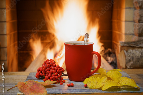 Red mug of tea, a bunch of mountain ash and yellow autumn leaves near a cozy fireplace, in a country house, winter or autumn holidays