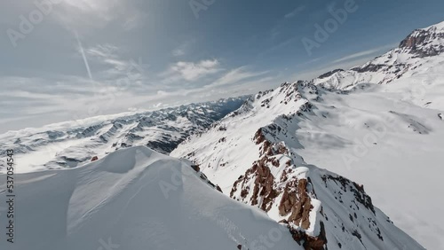 FPV sports drone flight alpine summit ridge natural rock texture snowy hilly valley with sunny sky picturesque landscape. Aerial view winter mountain epic scenery high cliff peak freeze glacier slope