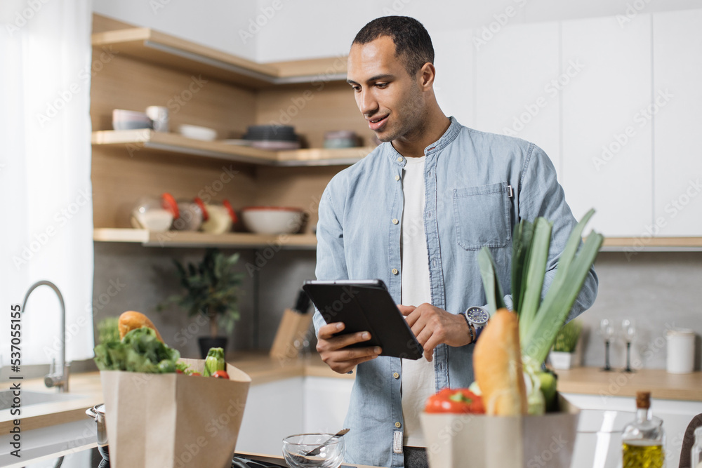 Attractive young arab man looking at an online recipe on a digital tablet. Handsome arabian male surfing the internet for recipes while making vegetarian salad in his modern kitchen. Attractive young arab man looking at an online recipe on a digital tablet. Handsome arabian male surfing the internet for recipes while making vegetarian salad in his modern kitchen.