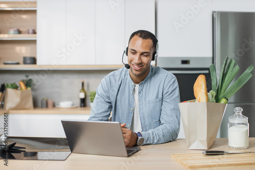 Young arab man, chef sitting in well-equipped modern kitchen having online video conversation with subscribers during vlog session how to cook delicious salad for breakfast looking at laptop .