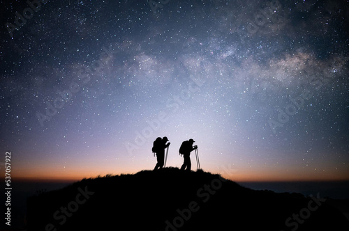 Silhouette of young couple hiker were standing at the top of the mountain looking at the stars and Milky Way over the twilight sky. Both of them were happy and free to travel.