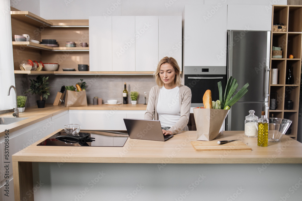 Cheerful young woman looking at an online recipe on a digital laptop pc. Attractive caucasian female surfing the internet for recipes while making vegetarian salad in her modern bright kitchen.