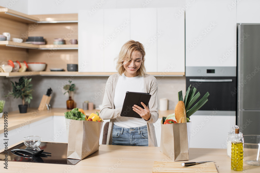 Positive cheerful young blond woman, searching menu recipe of healthy vegetarian vegetable salad in digital tablet in modern bright kitchen at home. Smiling female, food and technology concept.