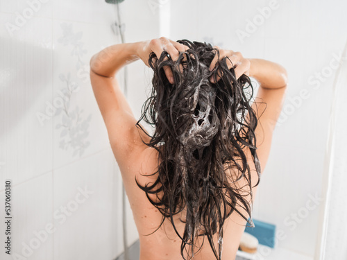 Naked woman with long hair takes a shower. Woman washes her hair with shampoo. Morning routine in modern urban apartment. White bathroom.