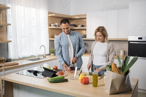 Young attractive couple in love preparing salad from fresh vegetables. Handsome sporty man and blond charming woman cooking dinner together and having fun in a new modern apartment.