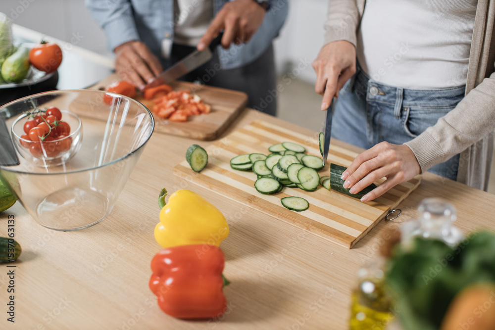 Close up of hands cutting fresh vegetables tomato and cucumber with knife, couple preparing salad. Unrecognizable man and woman cooking dinner together and having fun in a new modern apartment.