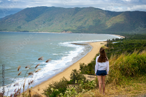 beautiful girl in white shirt stands on top of mountain overlooking tropical beach, holiday in northern queensland, cairns, australia, tropical paradise beach in australia © Jakub