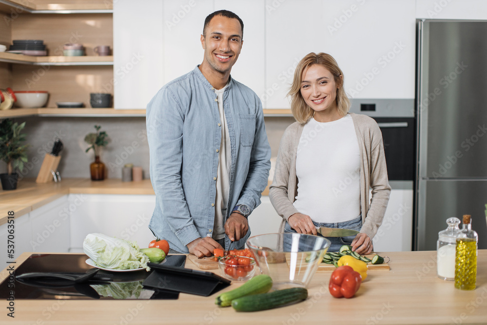 Young attractive couple in love preparing salad from fresh vegetables. Handsome sporty man and blond charming woman cooking dinner together and having fun looking at camera in a new modern apartment.