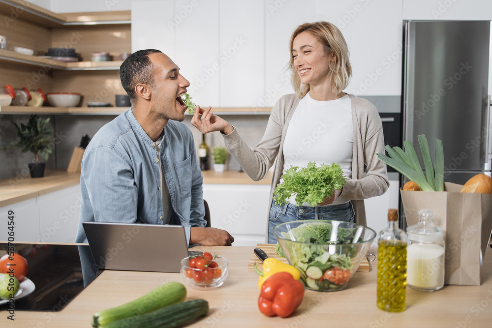 Cheerful married multinational couple using laptop while cooking healthy food in kitchen, blond young wife spouses having fun while feeding her husband with lettuce salad, preparing vegetable meal . Cheerful married multinational couple using laptop while cooking healthy food in kitchen, blond young wife spouses having fun while feeding her husband with lettuce salad, preparing vegetable meal .