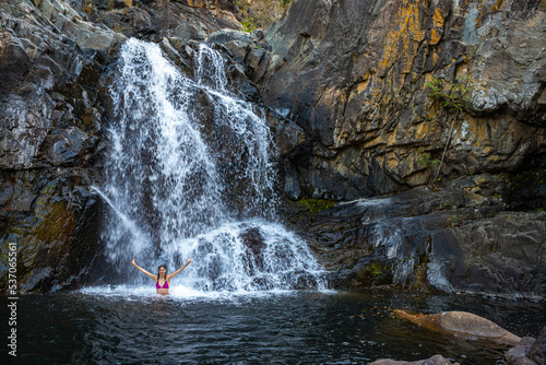 a beautiful girl in a pink bikini stands under a waterfall in a pool surrounded by massive rocks in jourama falls; vacation in paluma range national park in queensland, australia; cascading waterfalls photo