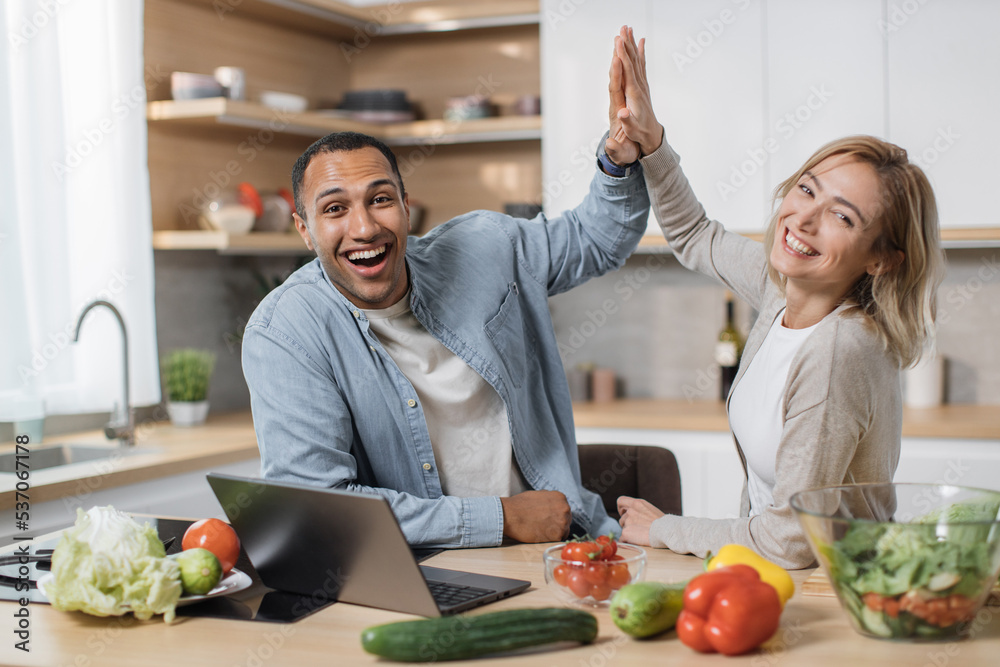 Attractive young multiethnic couple using laptop computer while preparing together healthy food diet vegetable salad at modern light kitchen giving high five at home .