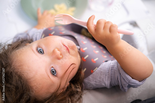 Top view portrait caucasian baby girl about 2 years old in bib eating pasta from plate sitting high chair, self feeding for kids, baby-led weaning idea, healthy nutrition of solid food for infant