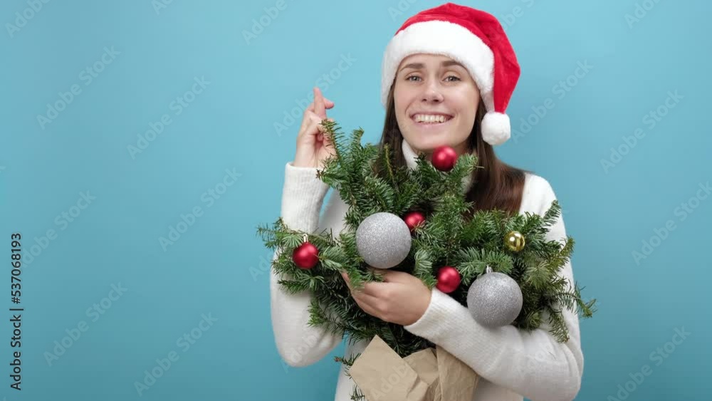 Pretty young Santa girl in Christmas hat hold bouquet of spruce branches keeping fingers crossed eyes closed making wish, isolated on blue background. Happy New Year celebration holiday party concept Pretty young Santa girl in Christmas hat hold bouquet of spruce branches keeping fingers crossed eyes closed making wish, isolated on blue background. Happy New Year celebration holiday party concept