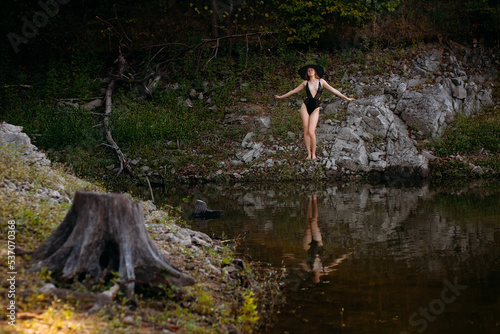 A young girl with long blond hair and a beautiful figure, in black openwork lingerie and a black wide hat, stands against the backdrop of a rock, on the banks of a picturesque river.