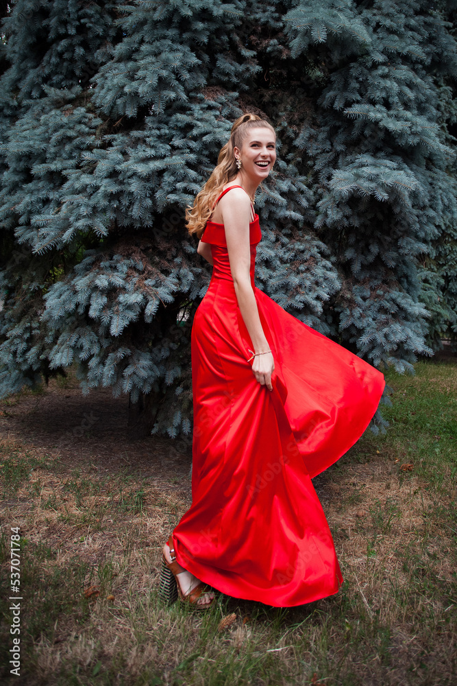 A cute smiling young girl with blond hair and beautiful makeup, in a long red dress, stands against the backdrop of trees in the park. A cute smiling young girl with blond hair and beautiful makeup, in a long red dress, stands against the backdrop of trees in the park.