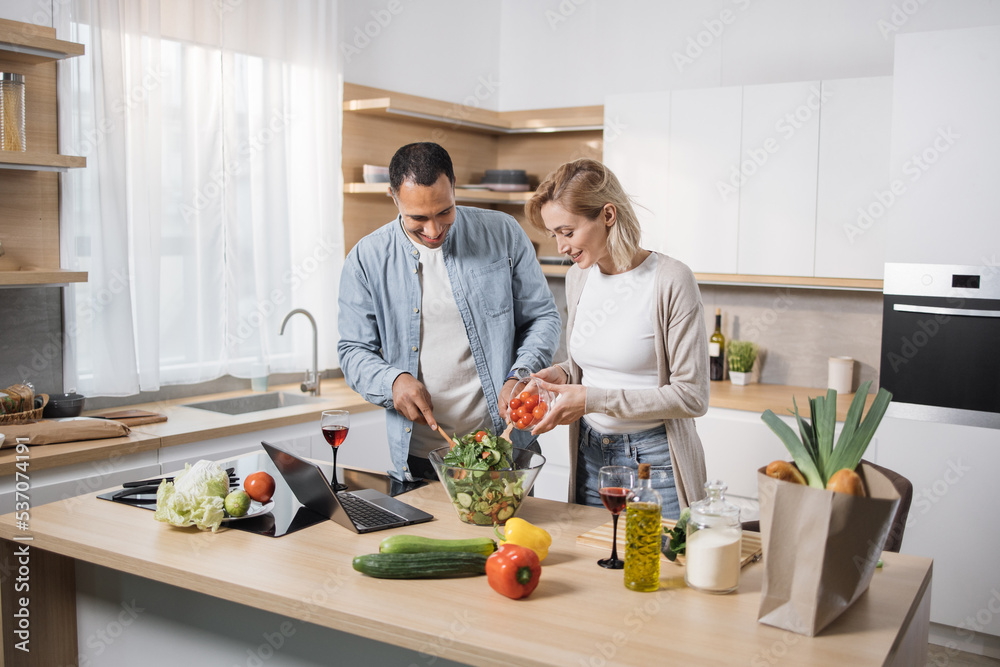 Young attractive couple in love preparing salad from fresh vegetables. Handsome sporty man mixing slices of ingredients and blond charming woman pouring olive oil into a glass bowl.