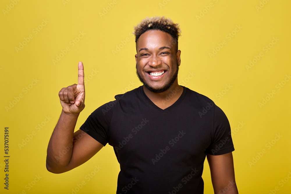 Joyful man poses in front of the camera in a black T-shirt in a yellow studio. On a yellow background stands a popular black guy with a smile on his face, pointing his finger up at the ceiling. Joyful man poses in front of the camera in a black T-shirt in a yellow studio. On a yellow background stands a popular black guy with a smile on his face, pointing his finger up at the ceiling.