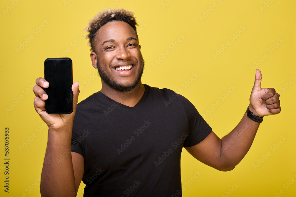 Muscular African-American shows the screen of a modern phone with a place for a layout. A satisfied black guy smiles and gives a thumbs up on a yellow background