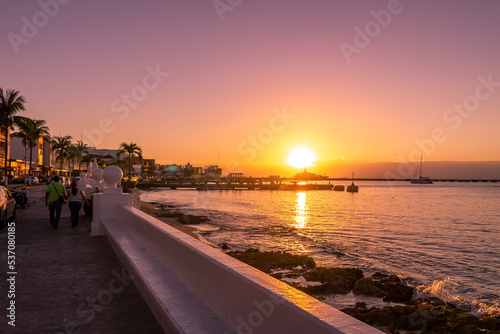 View of the waterfront of a tropical island during the golden hour. Palm trees and figures of people in the rays of the setting sun. On the horizon are the silhouettes of a pier and pleasure yachts. photo