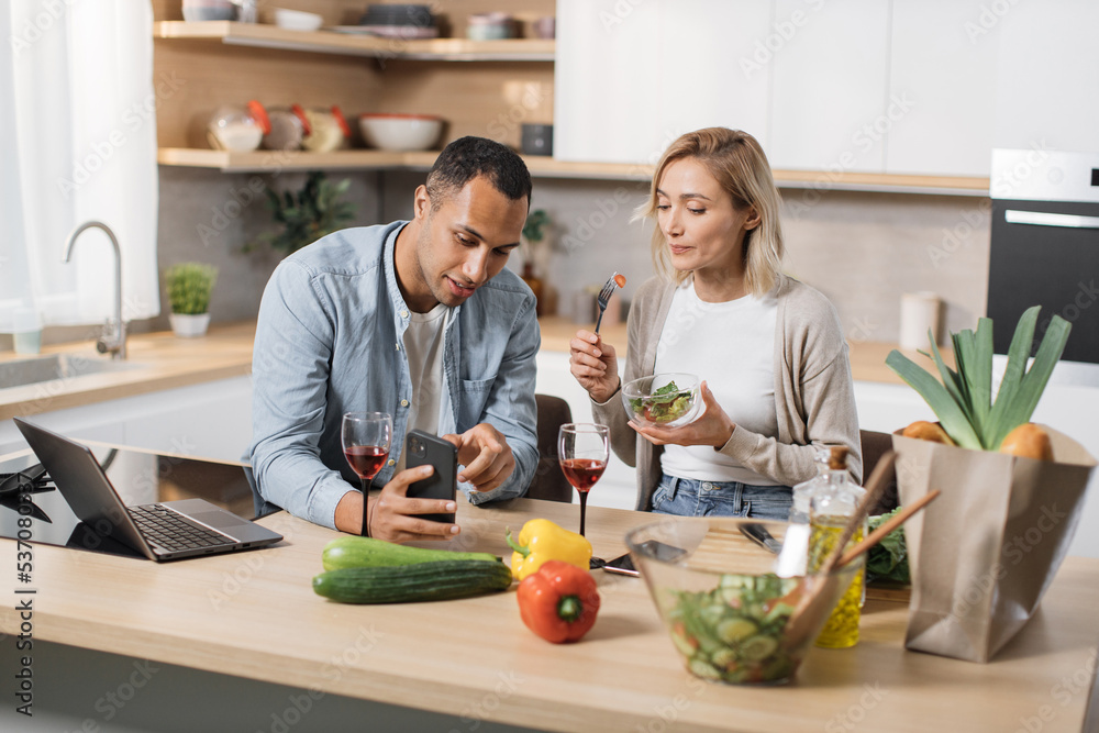 Young attractive couple in love eating salad from fresh vegetables using smartphone while having online video call with relatives or friend at modern kitchen at home.