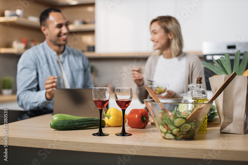 Focus on glasses with red wine. Cheerful couple using laptop eating diet salad from fresh organic vegetables and speaking to each other  smiling  relaxing after working day at modern kitchen at home.