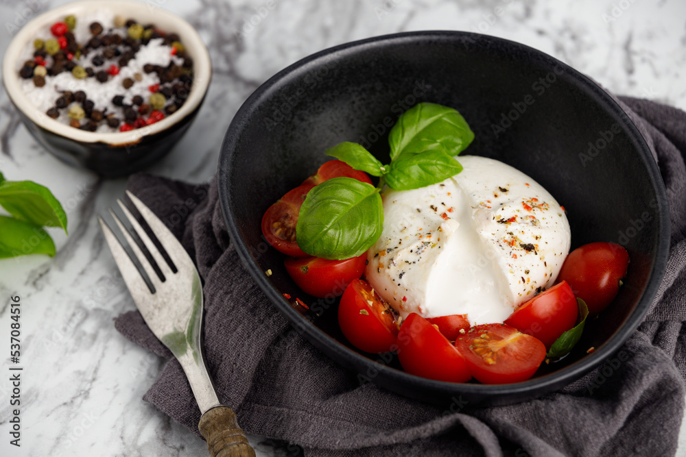Fresh Italian burrata with tomatoes, basil and spices on marble kitchen table, close up. Burrata cheese ball made from mozzarella and cream. Healthy food