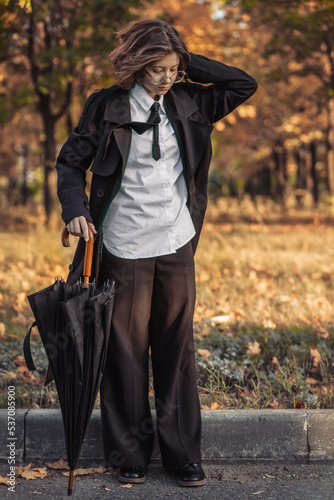 teenage girl with glasses, black raincoat with umbrella on asphalt road against the background of autumn forest, theme of subculture of dark academy, tinting, autumn background