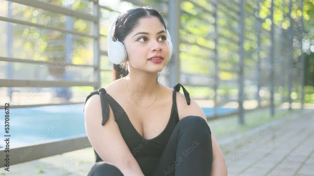 Attractive young indian woman lacing up sneakers and listening music on summer day in the stadium. Close-up shot of athlete preparing for training, running or exercising.