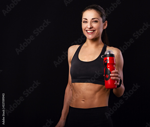 Happy pretty fit healthy laughing fitness woman in sporty top clothing holding sport flask and looking down on black background with empty copy space. Healthy lifestyle
