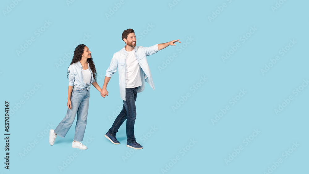 Smiling millennial caucasian man holding hand of arabic woman, walking, pointing finger to free space