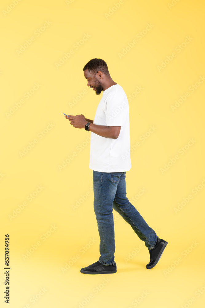 Black Guy Texting On Smartphone Using Application Walking, Yellow Background Black Guy Texting On Smartphone Using Application Walking, Yellow Background