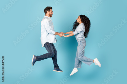 Smiling millennial caucasian male and arab lady holding hands, jump in air, enjoy romance on empty space