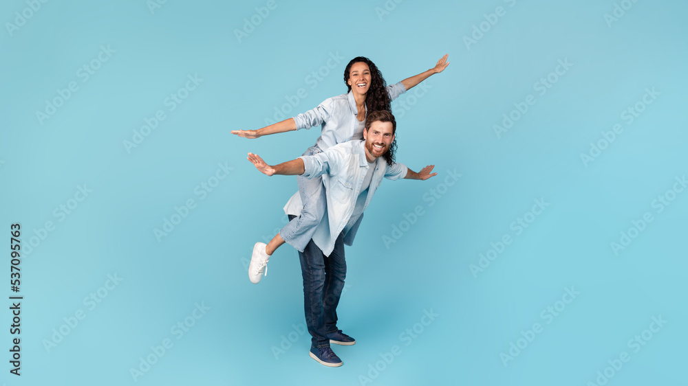 Cheerful millennial caucasian man holding middle eastern wife on his back, doing plane, have fun
