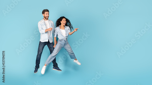 Laughing surprised excited millennial caucasian and arab couple jumping in air, showing finger to empty space