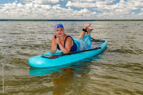 A woman with a mohawk in a pareo is sunbathing on a SUP board in a lake against the background of white clouds in a clear blue sky.