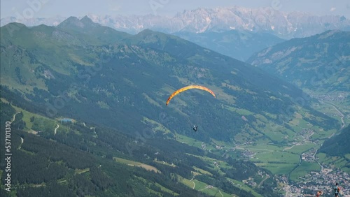 Man parachuting paragliding with a bright orange parachute slowly flying in the air sky in between green mountains with beautiful panorama view of the alps. Skydiving extreme action sport in summer.  (ID: 537101310)