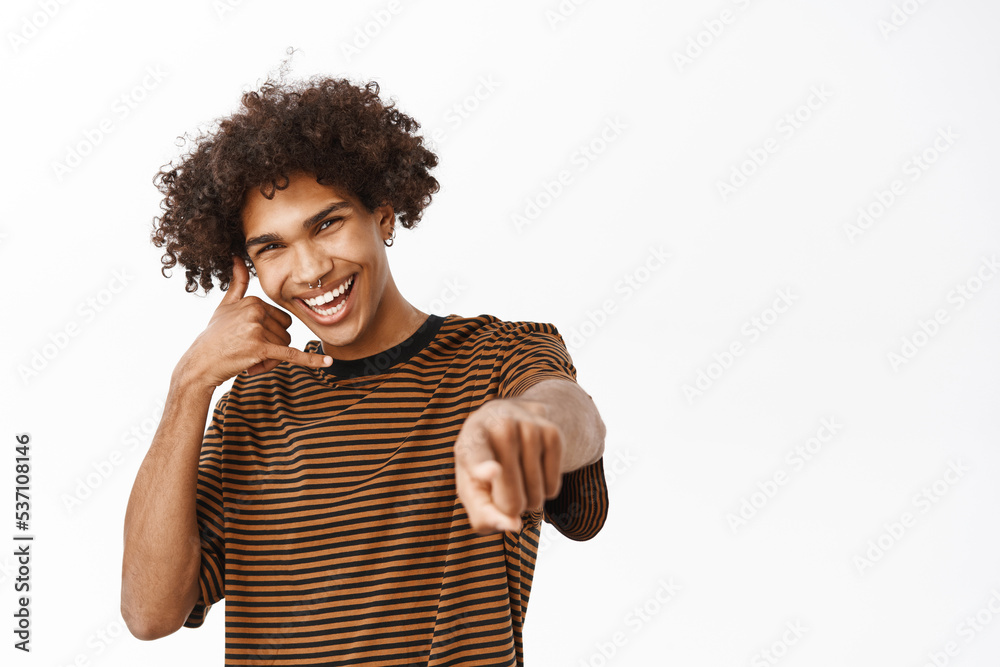 Call me. Handsome hispanic guy with curly hair, showing mobile phone near ear and pointing finger at camera, inviting you, standing over white background Call me. Handsome hispanic guy with curly hair, showing mobile phone near ear and pointing finger at camera, inviting you, standing over white background