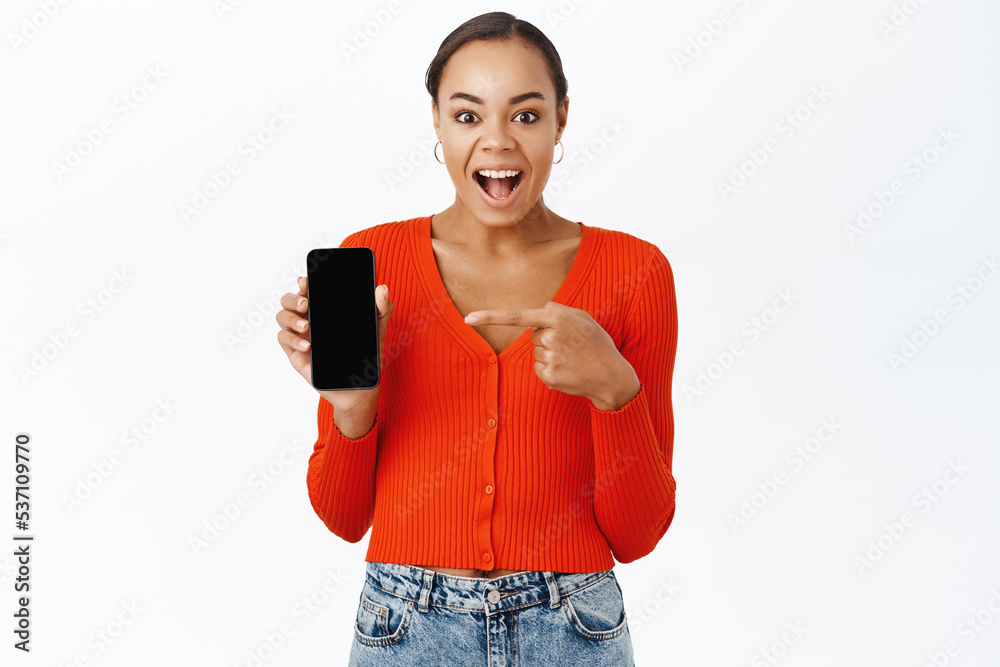 Portrait of woman pointing finger at mobile phone screen, showing smartphone application, standing over white background Portrait of woman pointing finger at mobile phone screen, showing smartphone application, standing over white background