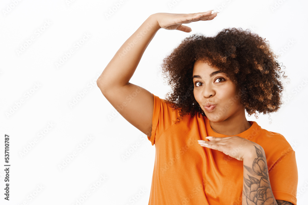 Portrait of cute and silly african american woman frames her head inside arms, pucker lips kissing, standing in orange tshirt over white background