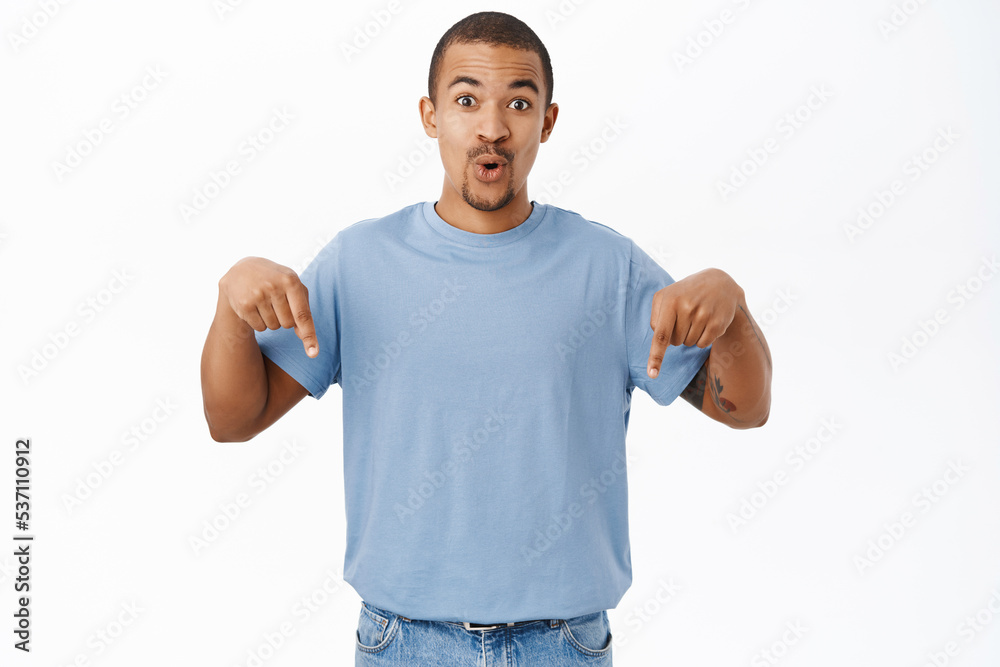 Surprised guy points fingers down and say wow, shows amazing news below, stands in blue tshirt over white background
